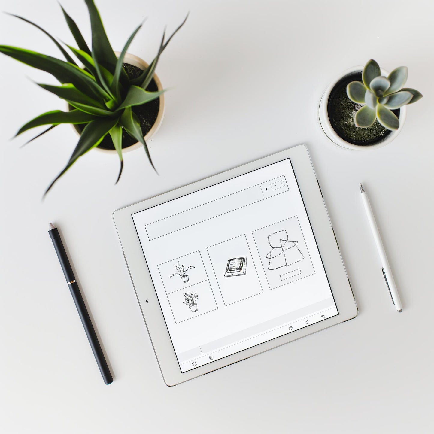 The Example product tablet displays simple line drawings on a white desk, flanked by two potted succulents, with a black pen to the left and a white stylus to the right.
