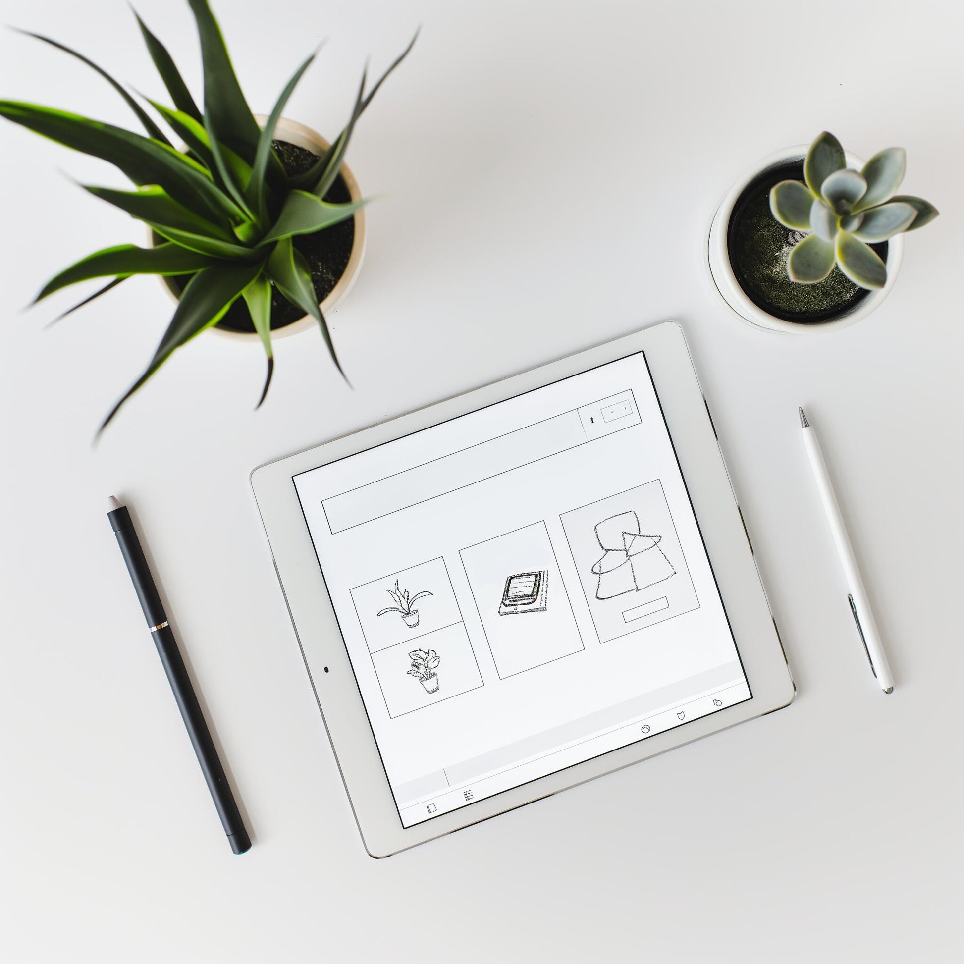 The Example product tablet displays simple line drawings on a white desk, flanked by two potted succulents, with a black pen to the left and a white stylus to the right.